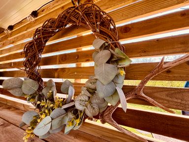 A decorative wreath with green leaves and yellow flowers hanging on a wooden wall with antlers and string lights at Yes Deer in Ashbourne