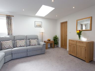 A living room with a grey sofa and patterned pillows next to wooden tables and a wooden cabinet with a mirror at Cornish Views in Camborne