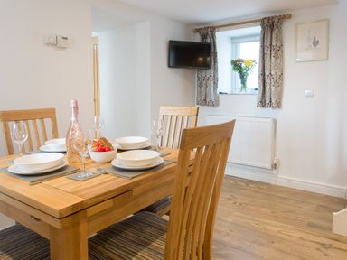 A dining area with a wooden table set for four near a window with curtains and a wall-mounted television at Cornish Views in Camborne