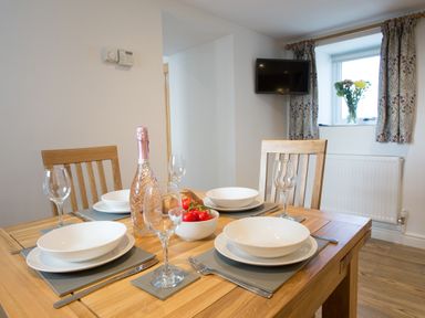 A dining room with a wooden table set with white plates wine glasses a bottle and a bowl of tomatoes at Cornish Views in Camborne