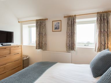 A bedroom with two windows with patterned curtains a wooden dresser with a television and a bed with gray pillows and blanket at Cornish Views Camborne