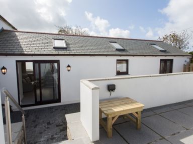 Exterior view of a white building with three skylights two lantern wall lights a wooden table and paved patio at Cornish Views in Camborne