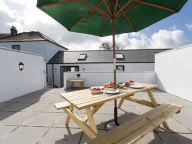 An outdoor patio with a wooden picnic table under a green umbrella set with plates and food at Cornish Views in Camborne