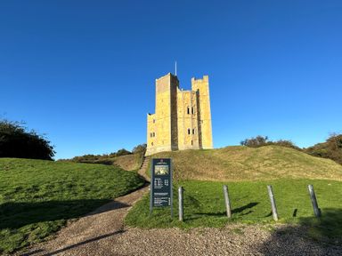 A stone castle on a grassy hill with a pathway and informational sign under a clear blue sky at Mundays in Orford