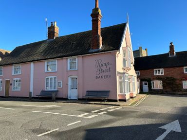 A street corner with a pink building labeled Pump Street Bakery and benches outside at Mundays in Orford