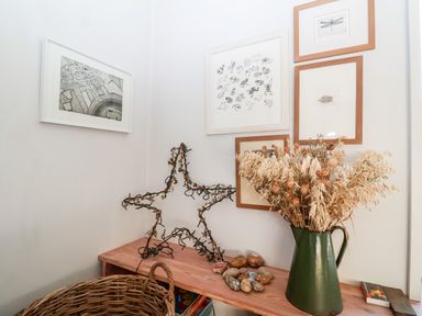 A corner with framed pictures on white walls a green jug with dried flowers a wooden shelf with stones and a star-shaped wire decoration at Mundays in Orford