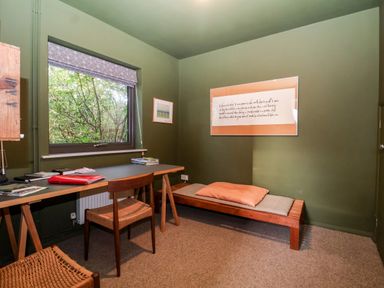 A room with a wooden desk and chairs a window with a patterned blind and a wooden daybed with cushions at Mundays Orford