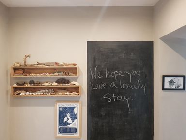 A wall with two wooden shelves holding rocks and driftwood a chalkboard with a handwritten message a framed map below the shelves and a framed drawing of a sheep on the right at Mundays Orford