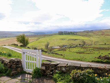 A white garden gate opening to a pathway with a stone wall and green fields with hills and trees in the background at Swinhope View in Hexham
