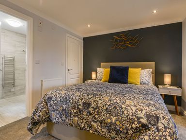 A bedroom with a bed with patterned bedding and cushions beside a door and an open doorway to a bathroom at Lilys Pad in Okehampton