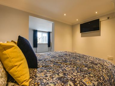 A bedroom with a bed covered in a floral bedspread yellow and dark blue pillows a window with dark curtains and a wall-mounted tv at Lilys Pad in Okehampton
