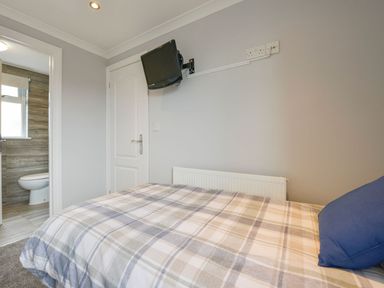 A bedroom with a bed with plaid bedding a blue pillow a wall mounted television a door and an open door leading to a bathroom at Lilys Pad in Okehampton
