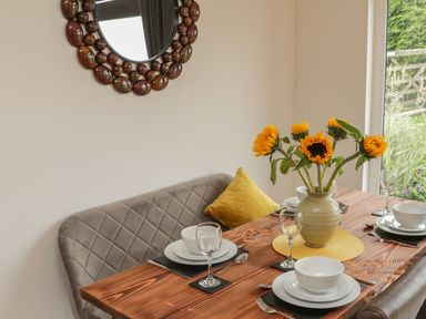 A dining area with a wooden table set with white dishes and wine glasses a vase with sunflowers and a cushioned bench with a yellow pillow at Windy Willows in Okehampton