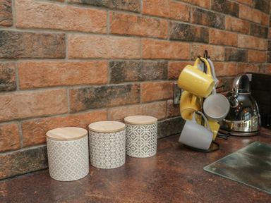 A kitchen counter with three patterned canisters wooden lids yellow and gray mugs on a rack and a stainless steel kettle at Windy Willows Okehampton