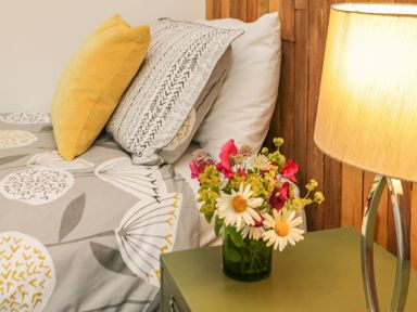 A bedside table with a lamp and a vase of flowers next to a bed with pillows and patterned bedding at Windy Willows in Okehampton