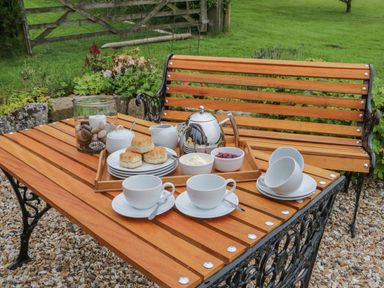 An outdoor wooden table and bench set with a tea tray holding scones jam cream a teapot and cups in a garden at Windy Willows in Okehampton