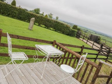 A wooden deck with white metal table and chairs overlooking a fenced grassy yard with hedges and a stone at Windy Willows in Okehampton
