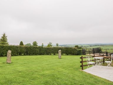 A grassy garden with two stone markers and white metal chairs and table on a wooden deck at Windy Willows in Okehampton