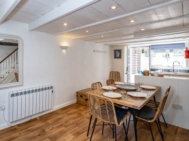 A dining area with a wooden table set for four and wicker chairs next to a kitchen counter with a window at Sea Horses in Port Isaac