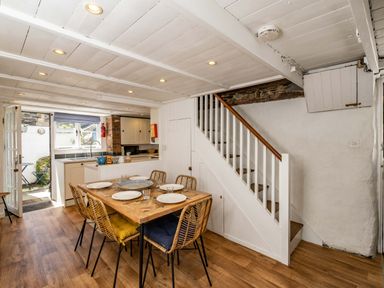 A dining area with a wooden table and six chairs next to a staircase and an open kitchen with a door leading outside at Sea Horses in Port Isaac