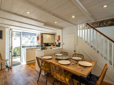 A dining area with a wooden table set with plates and glasses next to a kitchen and a staircase in Sea Horses in Port Isaac