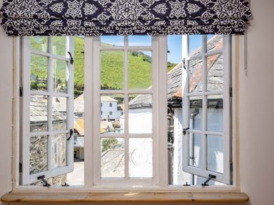 A view through an open window with a patterned curtain showing rooftops and green hills outside at Sea Horses in Port Isaac