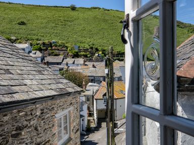 View of stone and slate roof buildings on a narrow street with a green hillside in the background seen through a window at Sea Horses in Port Isaac