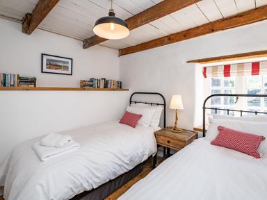 A bedroom with two single beds a wooden nightstand a lamp books on a shelf and a window with striped blinds at Sea Horses in Port Isaac