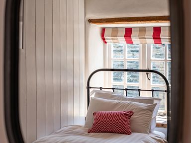 A bed with white pillows and a red cushion in front of a window with striped red and beige blinds at Sea Horses in Port Isaac