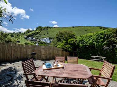 An outdoor patio with a wooden table and chairs with a tea set on the table and a green hill with houses in the background at Sea Horses in Port Isaac