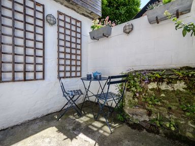 A small outdoor patio with a round table and two chairs next to white walls with wall lights and flower boxes at Sea Horses in Port Isaac