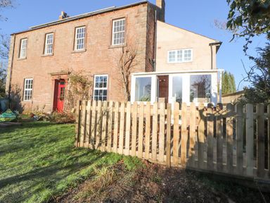 A two-story brick house with white windows a red door a glass porch and a wooden fence in a garden at Hillside Hideaway in Wigton