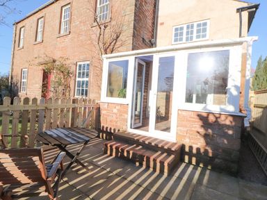 An outdoor patio with wooden table and chairs next to a brick house with a white framed glass door at Hillside Hideaway in Wigton