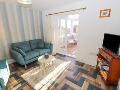A living room with a teal sofa a small round table a television on a wooden stand and sliding glass doors leading to a sunroom at Hillside Hideaway in Wigton