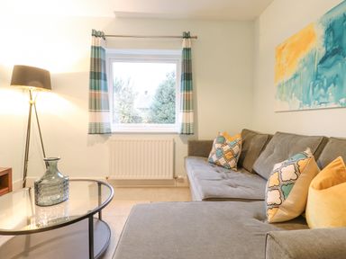 A living room with a gray sectional sofa with patterned and yellow pillows a glass coffee table with a vase a floor lamp and a window with striped curtains at Tandarra in Keswick