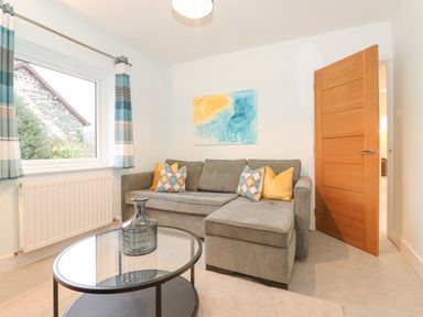 A living room with a grey sectional sofa with cushions a glass coffee table with a vase a window with striped curtains and an open wooden door at Tandarra in Keswick