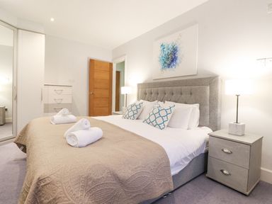A bedroom with a bed with rolled towels on the blanket two bedside tables with lamps a chest of drawers a wardrobe with a mirrored door a wooden door and artwork above the bed at Tandarra in Keswick