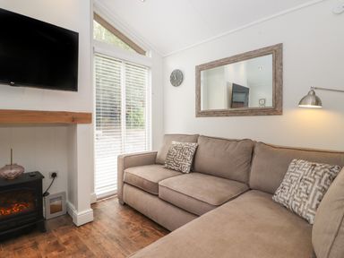 A living room with a beige sectional sofa a wall clock a mounted television and a window with blinds at Snuggledown Troutbeck 5 White Cross Bay near Windermere
