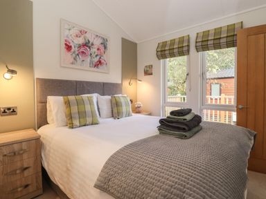 A bedroom with a double bed, bedside tables, folded towels on the bed, a painting above the bed, windows with blinds, and a wooden door at Snuggledown Troutbeck 5 White Cross Bay near Windermere