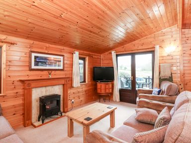 A wooden living room with a fireplace TV sofa armchairs wooden table and glass doors at Latrigg Lodge Number 9 Burnside Park near Keswick