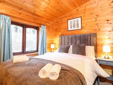 A bedroom with a bed with grey headboard and pillows with a brown blanket and rolled towels on it and a window with blue curtains at Latrigg Lodge Number 9 Burnside Park near Keswick