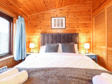 A bedroom with a double bed two bedside tables with lamps a window with blue curtains and wooden walls at Latrigg Lodge Number 9 Burnside Park near Keswick