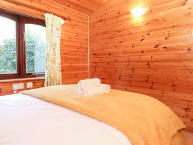 A bedroom with a bed covered by a beige blanket and white towels stacked on top a wooden wall and window with curtains at Latrigg Lodge Number 9 Burnside Park near Keswick