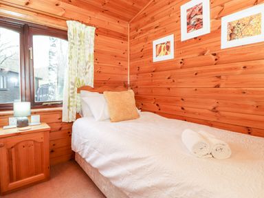 A bedroom with a single bed a wooden nightstand with a lamp a window with curtains and three framed pictures on wooden walls at Latrigg Lodge Number 9 Burnside Park near Keswick
