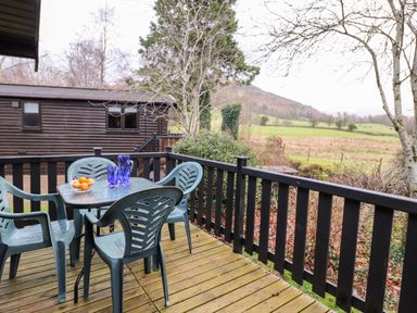 A wooden deck with a table and four chairs with blue glassware and fruit bowl overlooking fields at Latrigg Lodge Number 9 Burnside Park near Keswick