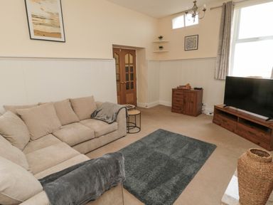 A living room with a beige sectional sofa a gray rug a wooden TV stand with a television a wooden cabinet corner shelves and framed pictures at Courtyard Cottage in Alnwick