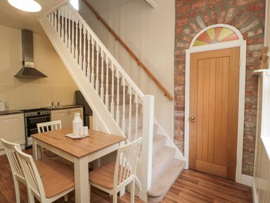 A kitchen dining area with a wooden table and four chairs next to carpeted stairs and a wooden door set in a brick wall at Courtyard Cottage in Alnwick
