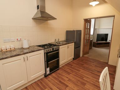 A kitchen with stove sink refrigerator and kettle seen from the doorway into a room with television and wooden furniture at Courtyard Cottage in Alnwick