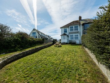A grassy backyard with a wooden picnic table and bench behind a two-story house at Tamarama House in Port Isaac