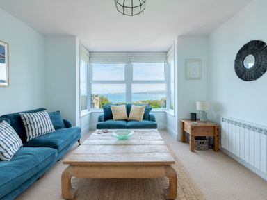 A living room with two blue sofas a wooden coffee table a side table with a lamp and a window with a sea view at Tamarama House in Port Isaac